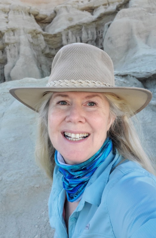 Trisha standing in the badlands wearing a sun hat and blue scarf, smiling at the camera