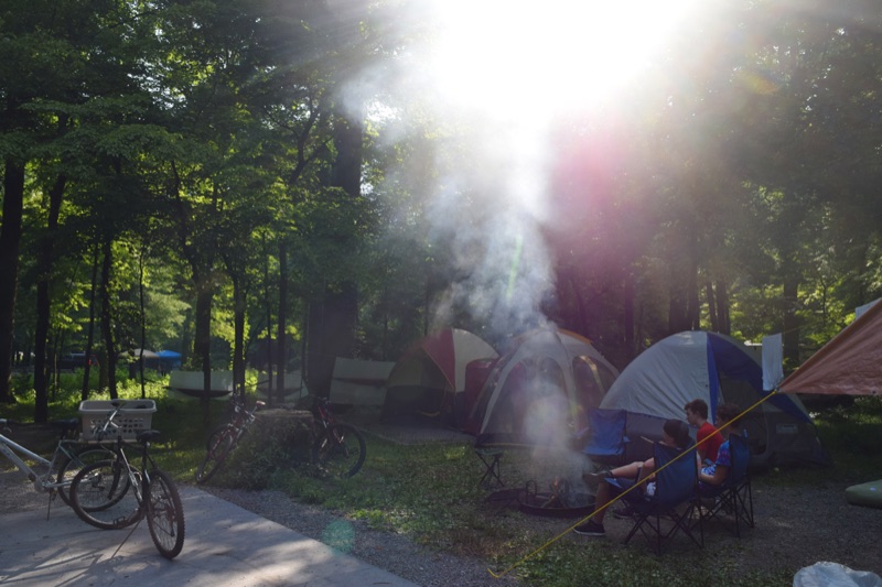 A shaded campsite with tents and bikes, campfire smoke catching the low sun, family members relaxing in camp chairs