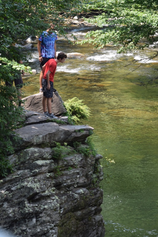 Two boys standing on a rock ledge above a green river pool, leaning over to look at the water below