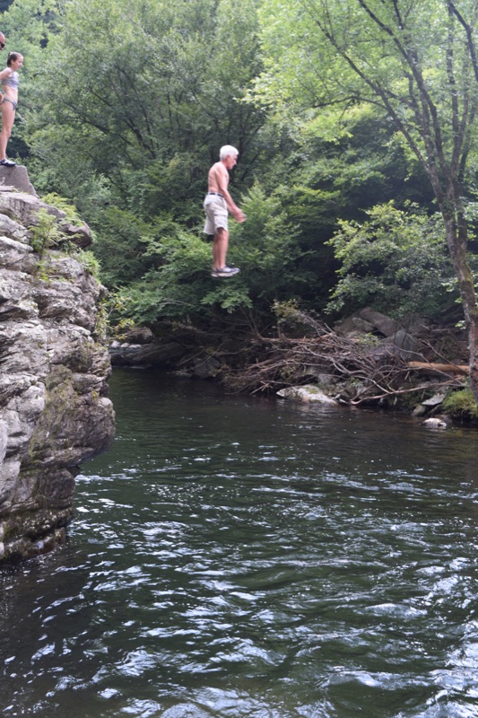 Randy caught mid-air, jumping feet-first off the ledge toward the river pool, a girl watching from the rock above