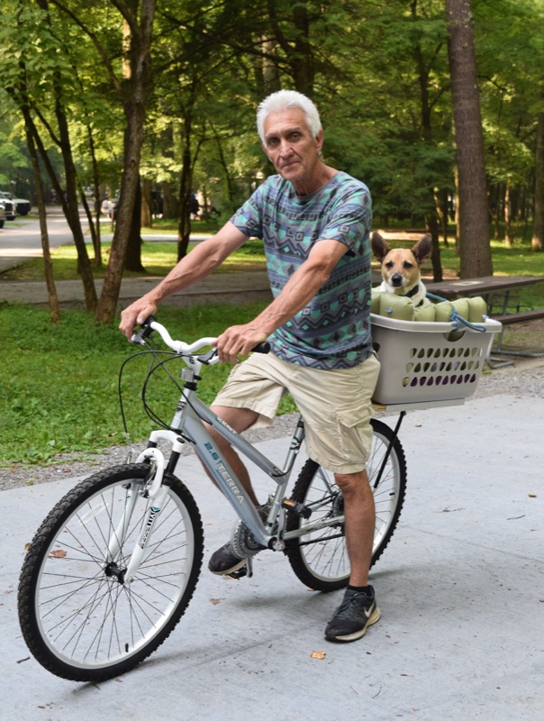 Randy on a bicycle on the campground road, the dog Pepper riding along in the front basket, green forest behind them