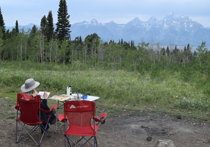 Trisha painting outdoors in the green sagebrush flats, brush in hand, the blue Tetons rising behind her