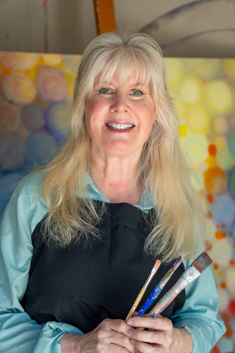Trisha Selgrath in her studio, holding paint brushes, in front of a bright abstract canvas