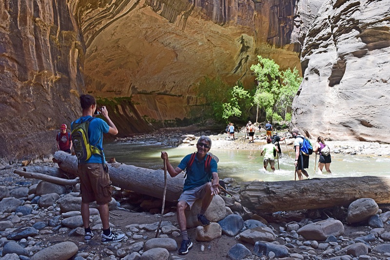 Hikers crossing the river beneath an immense overhanging sandstone wall in the Narrows