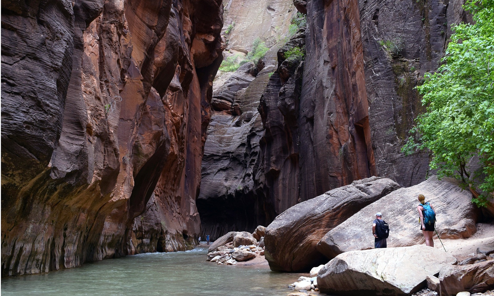 The Narrows — two hikers stand among boulders at the bend of a deep slot canyon in Zion National Park, sandstone walls rising around them